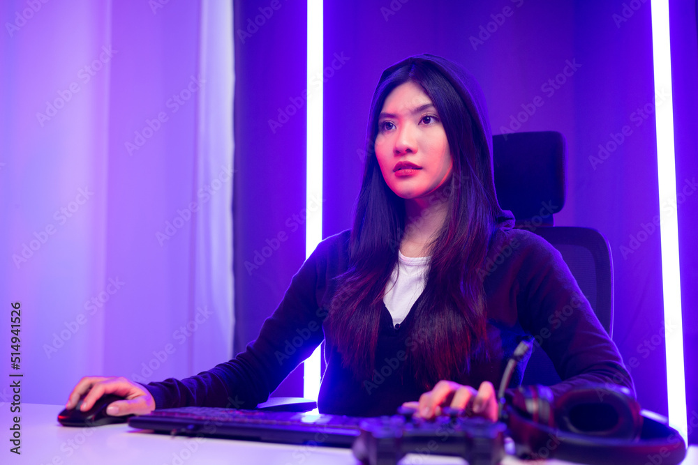 Playing video game. Young asian pretty woman sitting on chair with computer pc in living room. Happy female Professional Streamer chinese wearing hoodie playing game online in dark room neon light.
