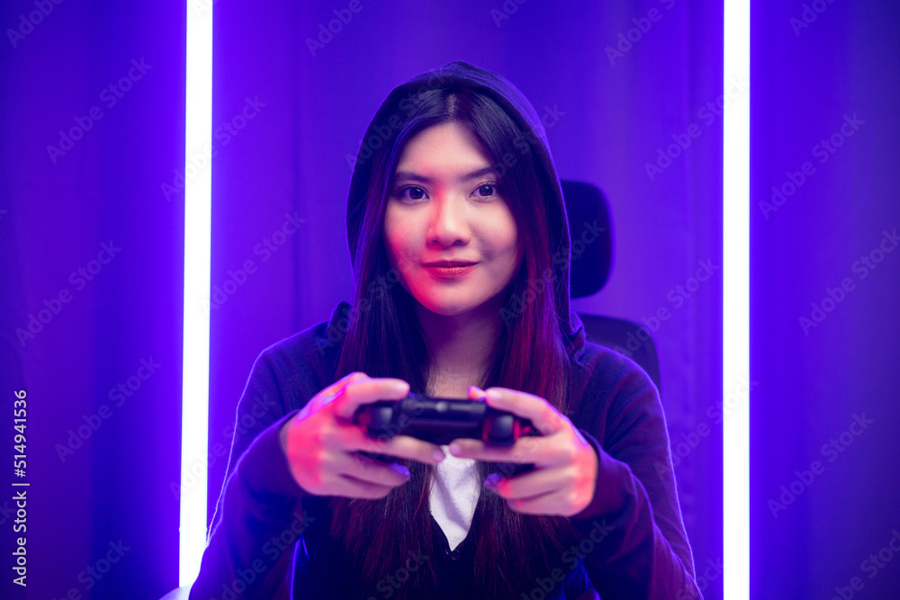 Playing video game. Young asian pretty woman sitting on chair holding joystick in living room. Happy female Professional Streamer chinese wearing hoodie playing game online in dark room neon light. Playing video game. Young asian pretty woman sitting on chair holding joystick in living room. Happy female Professional Streamer chinese wearing hoodie playing game online in dark room neon light.