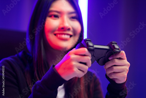 Playing video game. Young asian pretty woman sitting on chair holding joystick in living room. Happy female Professional Streamer chinese wearing hoodie playing game online in dark room neon light.