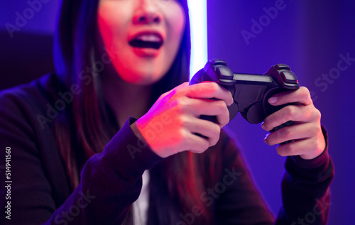 Playing video game. Young asian pretty woman sitting on chair holding joystick in living room. Happy female Professional Streamer chinese wearing hoodie playing game online in dark room neon light.