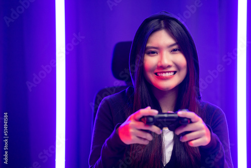 Playing video game. Young asian pretty woman sitting on chair holding joystick in living room. Happy female Professional Streamer chinese wearing hoodie playing game online in dark room neon light.
