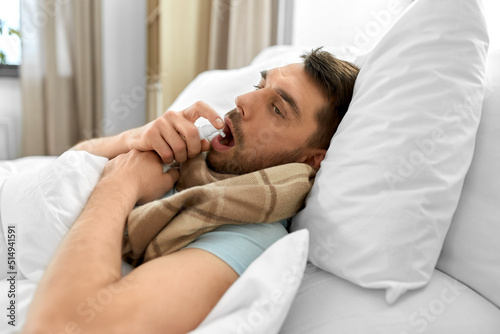 people and health problem concept - unhappy sick man spraying his throat with oral spray lying in bed at home