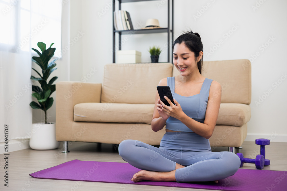 Lifestyle Attractive asian young fitness woman holding smartphone relaxing after workout at home. Smiling female using cell phone checking newsfeed on social media while exercise