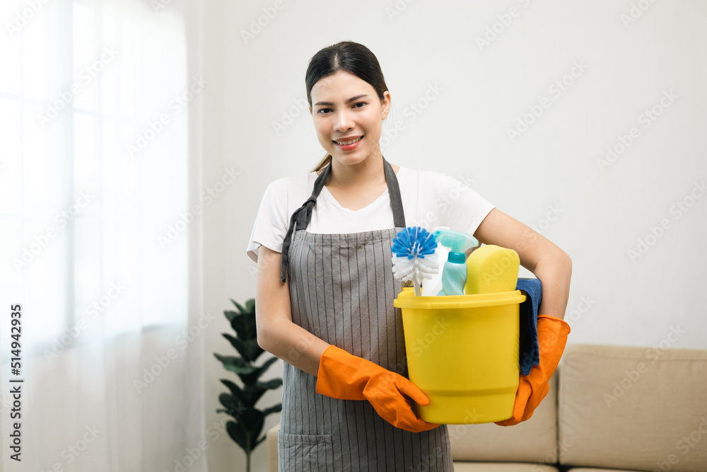 Young housekeeper woman holding bucket of cleaning products ready for cleaning home background.