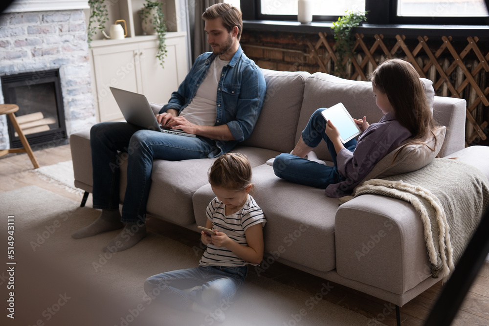 Obraz premium Young family of three holding and using different electronic devices while sitting on sofa in the living room