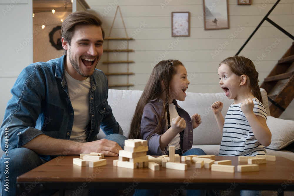 Joyful father with daughters plays at home. Kids remove wooden blocks from tower. Board game.
