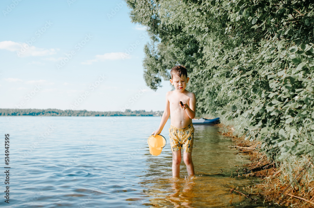 Fototapeta premium Child playing on lake with bucket and shell. Boy resting in nature during summer holidays.