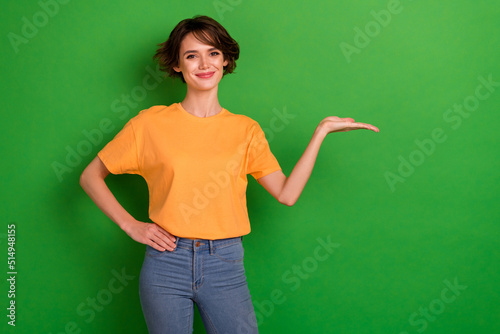 Photo of sweet funny young lady wear orange outfit smiling holding arm empty space isolated green color background