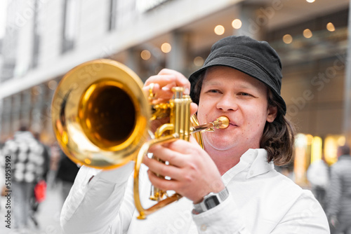 Young caucasian hally cheerful man in white shirt and hat playing funky jazz on golden trumpet with pleasure standing on crowded blurred downtown street close up. Musical lifestyle, hobby, education
