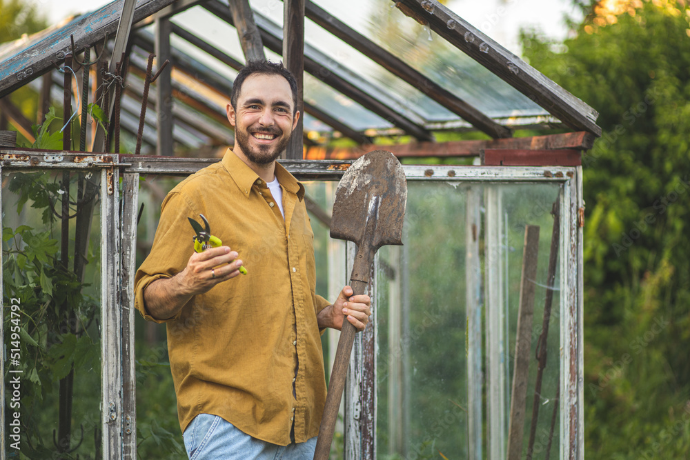 A man with a shovel and secateurs in his hands is sitting near a greenhouse. Farming. The process of caring for the greenhouse and seedlings. Rural life. Plant breeding and selection. A man with a shovel and secateurs in his hands is sitting near a greenhouse. Farming. The process of caring for the greenhouse and seedlings. Rural life. Plant breeding and selection.