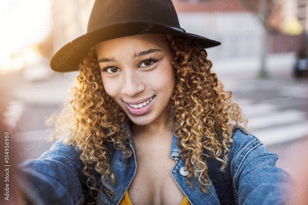Hispanic woman traveler with hat, young Hispanic backpacker tourist smiling happy making selfie next to camera in the city