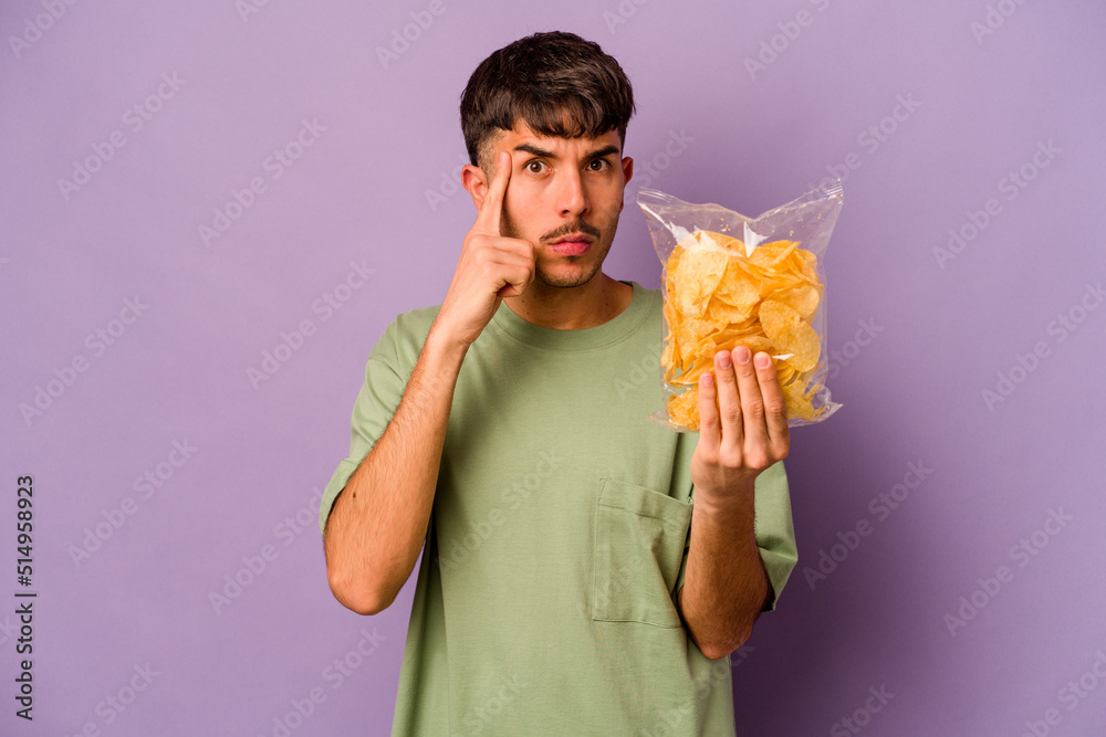 Young hispanic man holding crisps isolated on purple background pointing temple with finger, thinking, focused on a task. Young hispanic man holding crisps isolated on purple background pointing temple with finger, thinking, focused on a task.