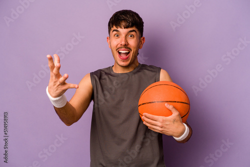 Young hispanic basketball player man isolated on purple background receiving a pleasant surprise, excited and raising hands.
