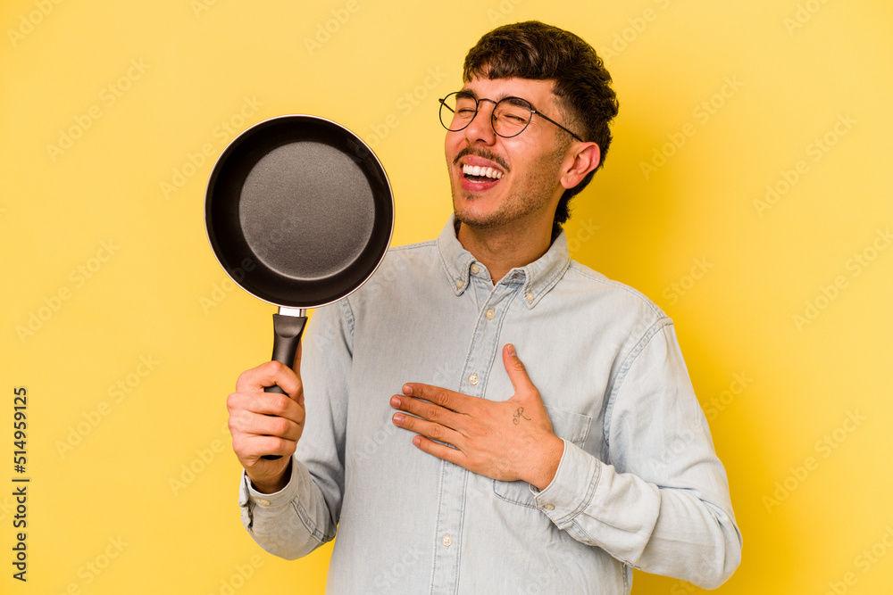 Young hispanic cooker holding frying pan isolated on yellow background laughs out loudly keeping hand on chest. Young hispanic cooker holding frying pan isolated on yellow background laughs out loudly keeping hand on chest.