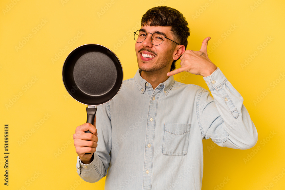 Young hispanic cooker holding frying pan isolated on yellow background showing a mobile phone call gesture with fingers. Young hispanic cooker holding frying pan isolated on yellow background showing a mobile phone call gesture with fingers.