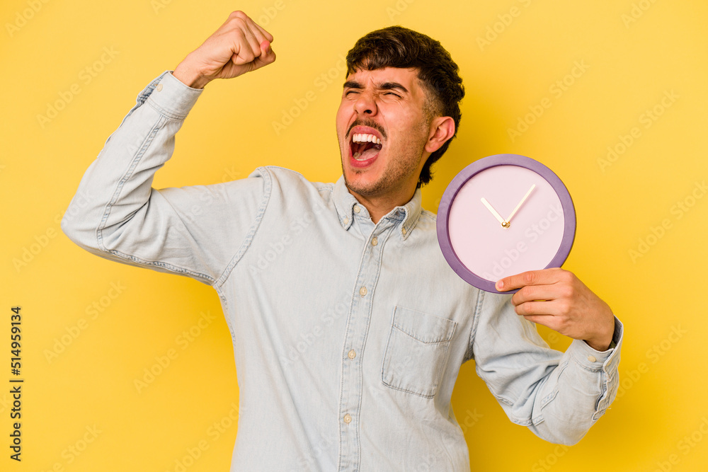 Young hispanic man holding a clock isolated on yellow background raising fist after a victory, winner concept.