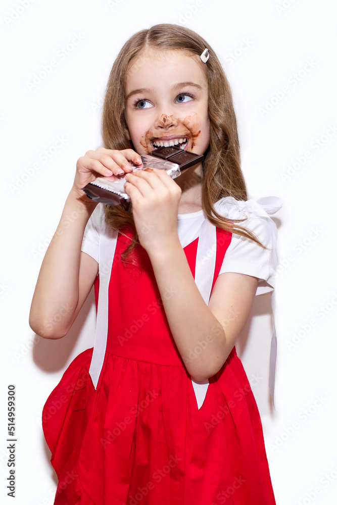 A little girl eats a bar of chocolate on a white vertical background. Happy child with face and hands covered in chocolate. The concept of children's happiness and love for sweets A little girl eats a bar of chocolate on a white vertical background. Happy child with face and hands covered in chocolate. The concept of children's happiness and love for sweets