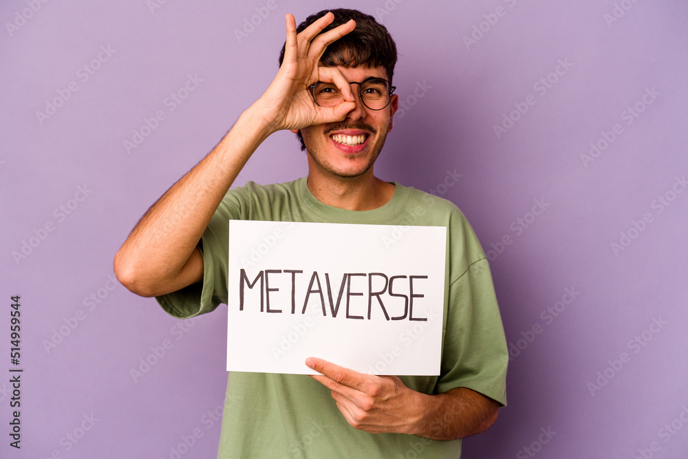 Young hispanic man holding meta verse placard isolated on purple background excited keeping ok gesture on eye. Young hispanic man holding meta verse placard isolated on purple background excited keeping ok gesture on eye.