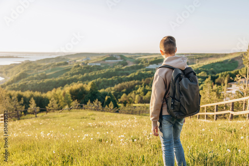 Tourist with backpack standing on top of hill in grass field and enjoying beautiful landscape view. Rear view of teenage boy hiker resting in nature. Active lifestyle. Concept of local travel © Lyubov