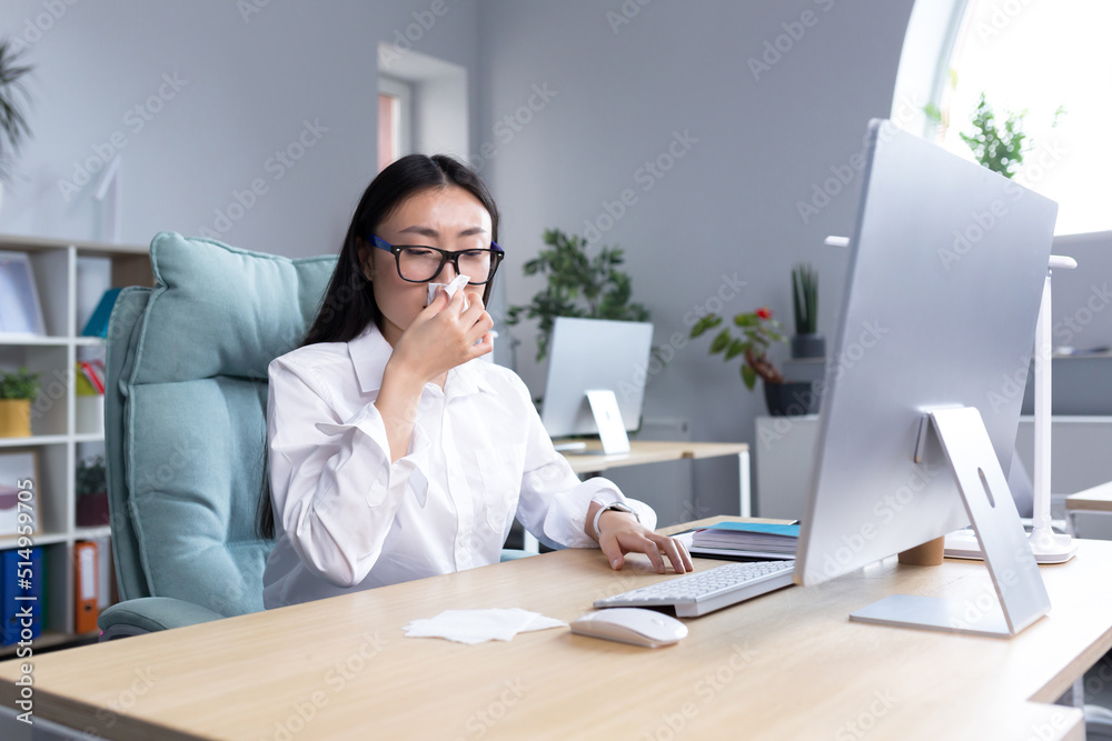 Sick Asian female office worker, has cold and flu, coughs and sneezes into tissue, works at computer. Sick Asian female office worker, has cold and flu, coughs and sneezes into tissue, works at computer.