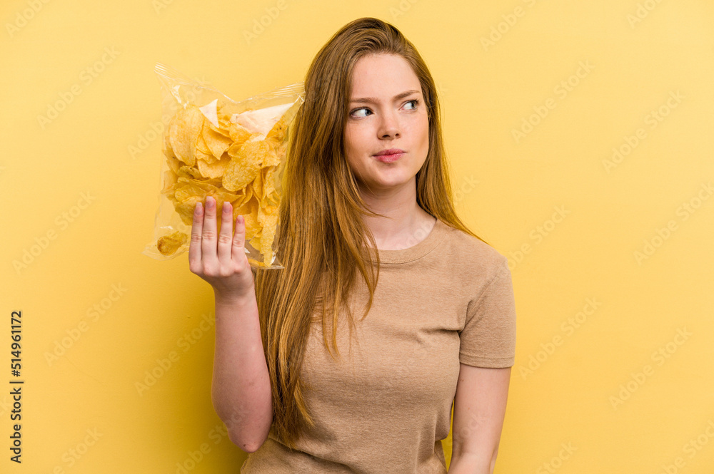 Young caucasian woman holding a bag of chips isolated on yellow background confused, feels doubtful and unsure. Young caucasian woman holding a bag of chips isolated on yellow background confused, feels doubtful and unsure.
