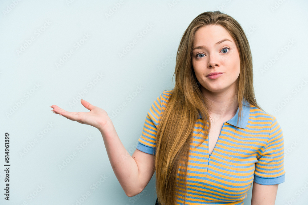 Young caucasian woman isolated on blue background showing a copy space on a palm and holding another hand on waist. Young caucasian woman isolated on blue background showing a copy space on a palm and holding another hand on waist.