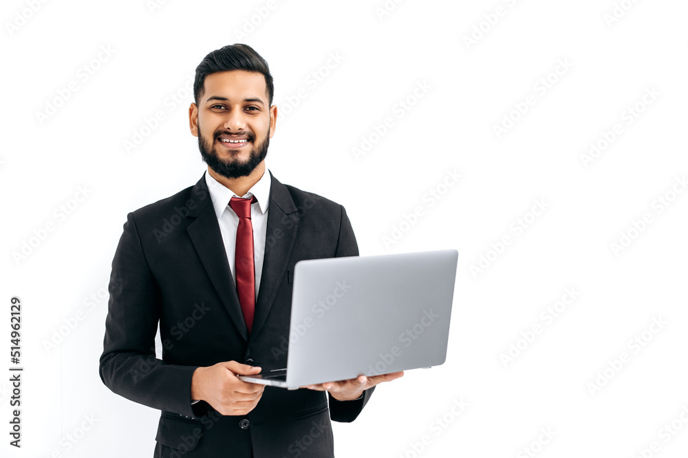 Successful handsome indian or arabian man in business suit, male entrepreneur, holding open laptop in hands, looks at camera, standing over isolated white background, smiles friendly. Copy-space