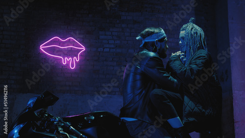 A girl in love and a guy are sitting on a super sport motorcycle flirting and hugging against the background of a neon sign