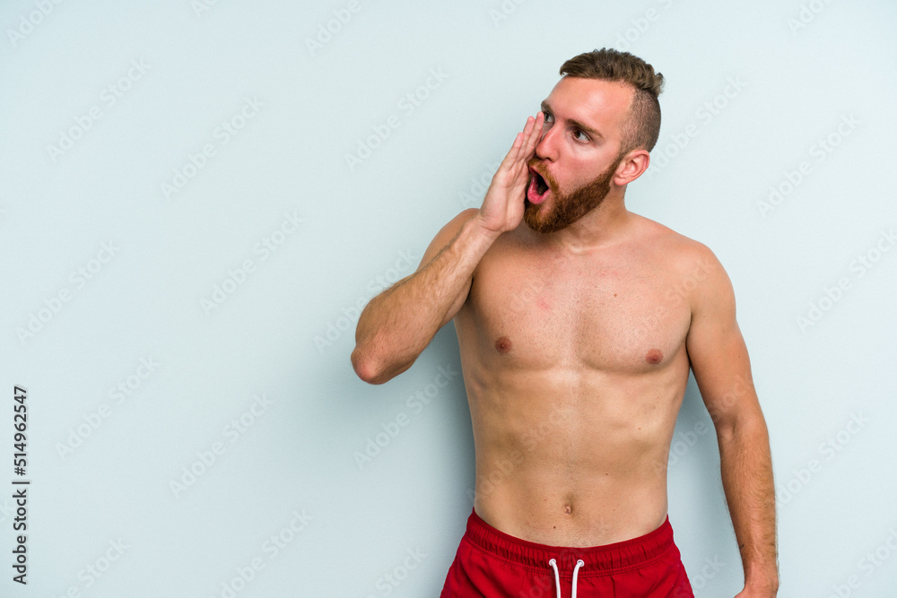 Young caucasian man wearing a swimsuit isolated on blue background is saying a secret hot braking news and looking aside