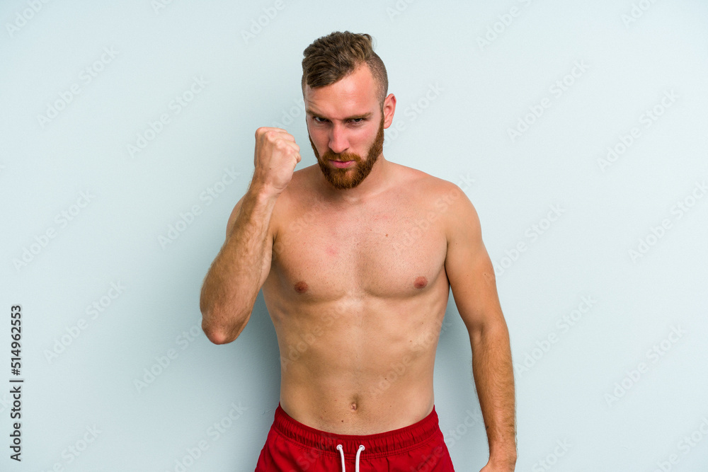 Young caucasian man wearing a swimsuit isolated on blue background showing fist to camera, aggressive facial expression.