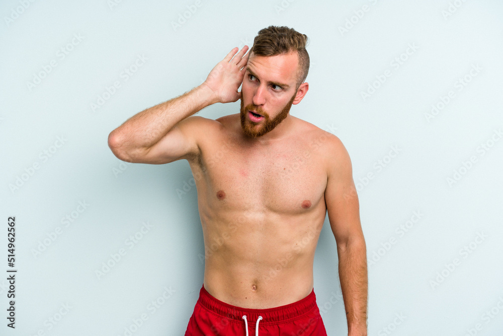 Young caucasian man wearing a swimsuit isolated on blue background trying to listening a gossip.