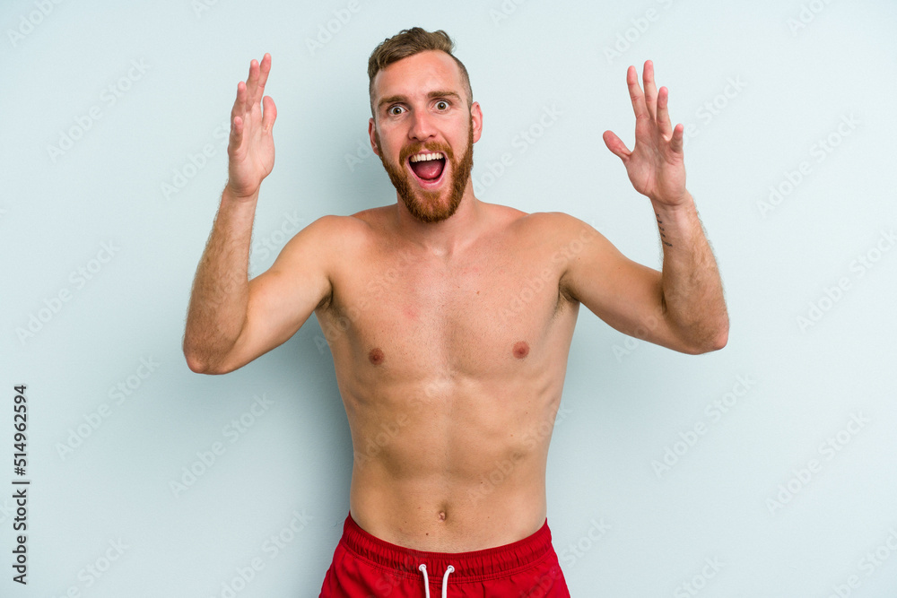 Young caucasian man wearing a swimsuit isolated on blue background receiving a pleasant surprise, excited and raising hands. Young caucasian man wearing a swimsuit isolated on blue background receiving a pleasant surprise, excited and raising hands.