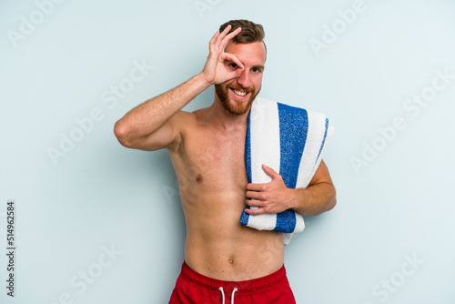 Young caucasian man going to the beach holding a towel isolated on blue background excited keeping ok gesture on eye.
