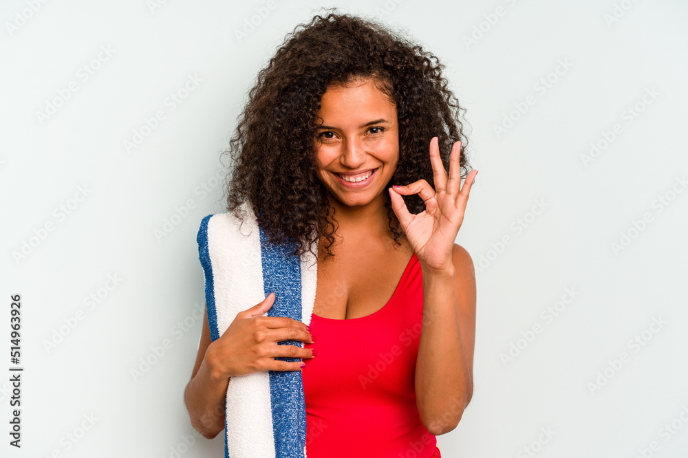 Young brazilian woman going to the beach holding a towel isolated on blue background cheerful and confident showing ok gesture. Young brazilian woman going to the beach holding a towel isolated on blue background cheerful and confident showing ok gesture.