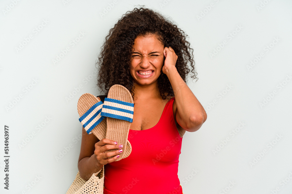 Young brazilian woman going to the beach holding a sandals isolated on blue background covering ears with hands.