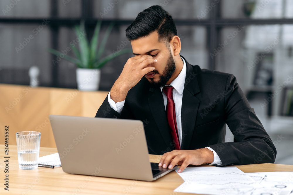 Tired exhausted indian or arabian businessman in a suit, sit at workplace in office, rubbing his nose bridge, feelings fatigue after long time work with laptop, overworked, need rest, coffee break Tired exhausted indian or arabian businessman in a suit, sit at workplace in office, rubbing his nose bridge, feelings fatigue after long time work with laptop, overworked, need rest, coffee break