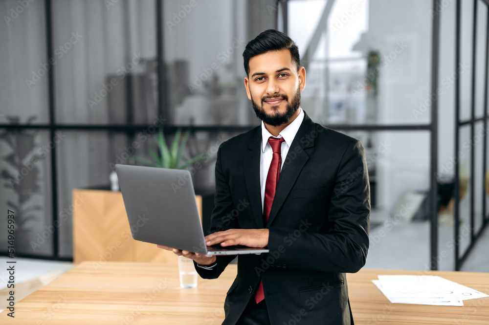 Attractive confident successful businessman of indian and arabian ethnicity, in a formal suit, ceo company, stands in his office near workplace, holdas an open laptop, looks at camera, smiles friendly Attractive confident successful businessman of indian and arabian ethnicity, in a formal suit, ceo company, stands in his office near workplace, holdas an open laptop, looks at camera, smiles friendly