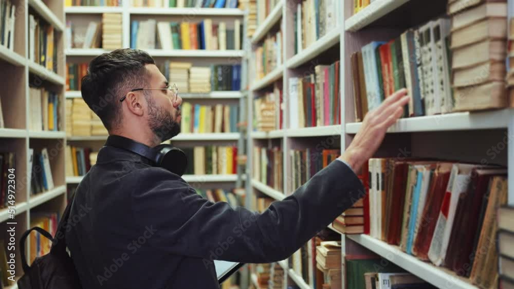 Young man with beard wearing eyeglasses and having headphones on neck standing by bookcase and choosing books, checking list of literature on tablet, student in library. Concept of education Young man with beard wearing eyeglasses and having headphones on neck standing by bookcase and choosing books, checking list of literature on tablet, student in library. Concept of education