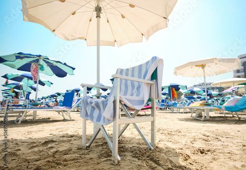 Fototapeta Naklejka Na Ścianę i Meble -  Chair with striped towel under an umbrella le at the beach on a sunny summer day. In the background, the beach equipped with sun loungers and umbrellas along the Adriatic Sea in Italy.