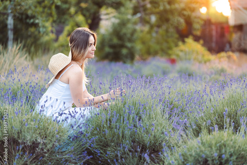 Photo of a caucasian young woman in a dress walking through a lavender field and picking flowers.