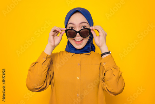 Shocked young Asian Muslim woman dressed in orange shirt looking at camera with hand touching his glasses  interested to big promo isolated on yellow background