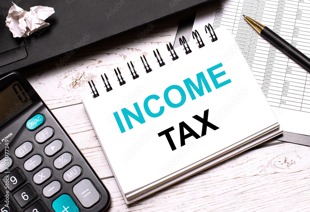 On a light-colored office table is a computer, a black calculator, a pen, and a notebook with the text INCOME TAX. Business concept. On a light-colored office table is a computer, a black calculator, a pen, and a notebook with the text INCOME TAX. Business concept.