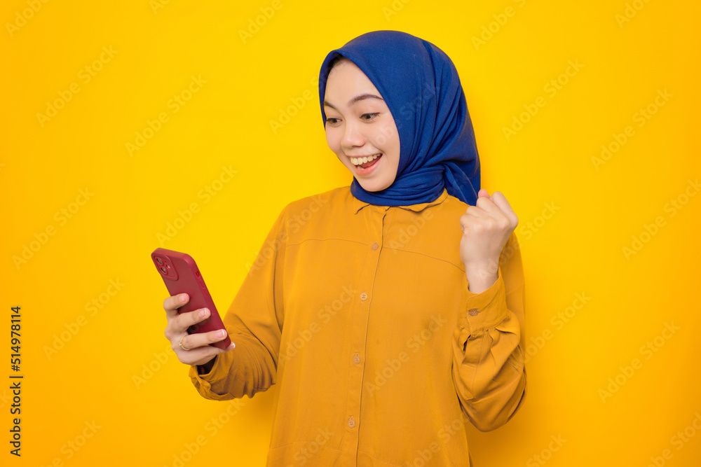 Excited young Asian Muslim woman dressed in orange shirt looking at smartphone getting good news, celebrating luck or success isolated on yellow background Excited young Asian Muslim woman dressed in orange shirt looking at smartphone getting good news, celebrating luck or success isolated on yellow background