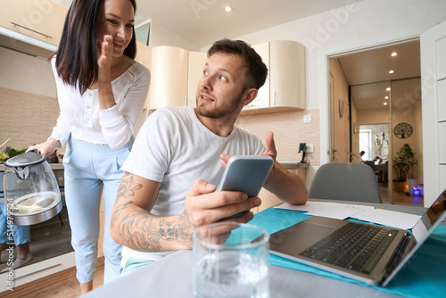 Wife giving advice to husband, working at laptop with smartphone