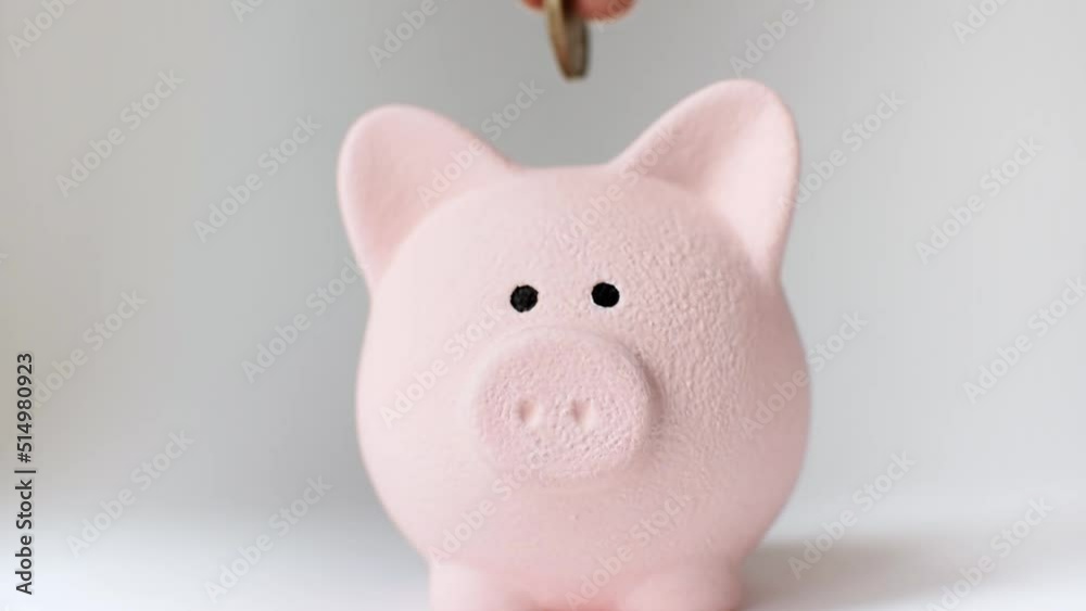 Close-up of a hand putting coins one by one into a pink piggy bank on a white background. Money box for savings. Economy, investment, future, finance, pension, dreams concept. Close-up of a hand putting coins one by one into a pink piggy bank on a white background. Money box for savings. Economy, investment, future, finance, pension, dreams concept.