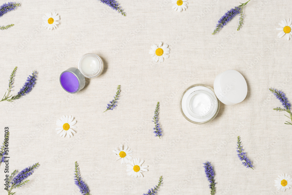 Cosmetic round containers with face cream and herbal balsam, purple veronica and white daisy wild flowers on natural textile table background. Natural organic beauty product concept. Flatlay