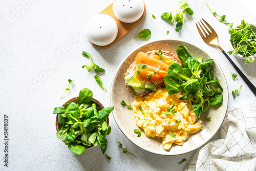Fotografia Scrambled eggs, sandwich with cream cheese and salmon and salad leaves at white stone table