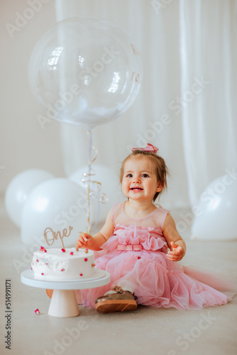 Cute one year girl in pink beautiful dress and cute little bow posing with white birthday cake and balloons in the studio. Party concept