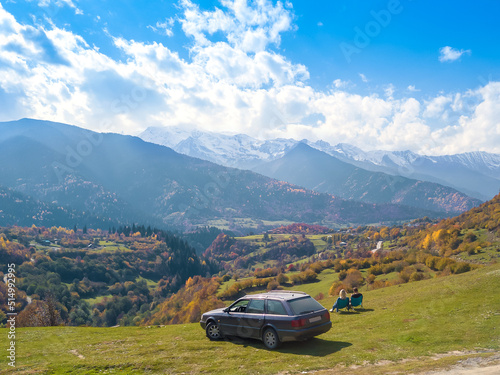 Man and a woman are sitting on chairs near the car and enjoying the amazing beautiful autumn mountain landscape of Svaneti, view from the drone. Concept of a tourist lifestyle, traveling by car.