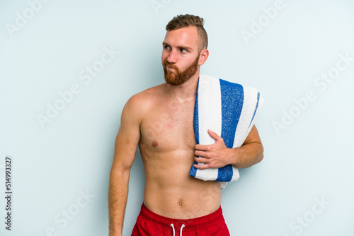 Young caucasian man going to the beach holding a towel isolated on blue background confused, feels doubtful and unsure.